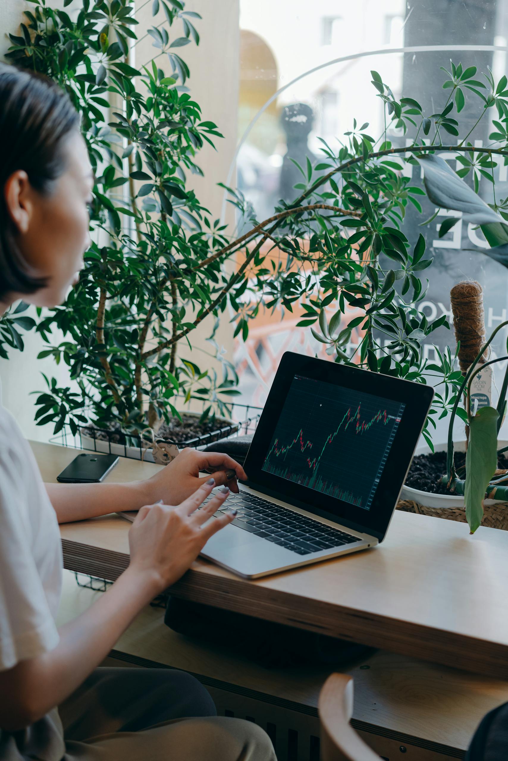 Businesswoman checks financial graphs on laptop in cozy café, surrounded by plants, showcasing remote work lifestyle.